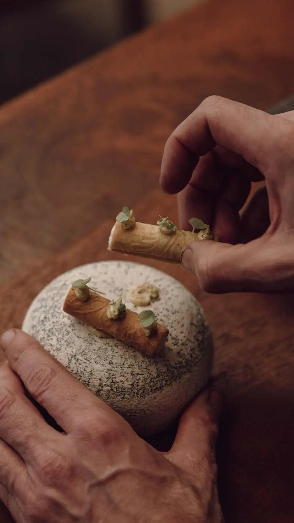 Mãos servindo um prato do menu degustação sobre a mesa de madeira.
