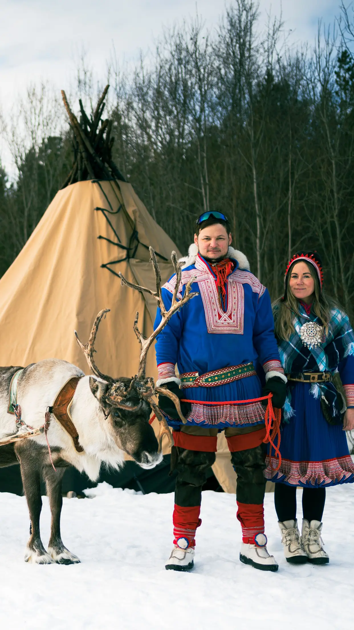 Anfitriões sámi em trajes tradicionais diante do lavvu, com uma rena.